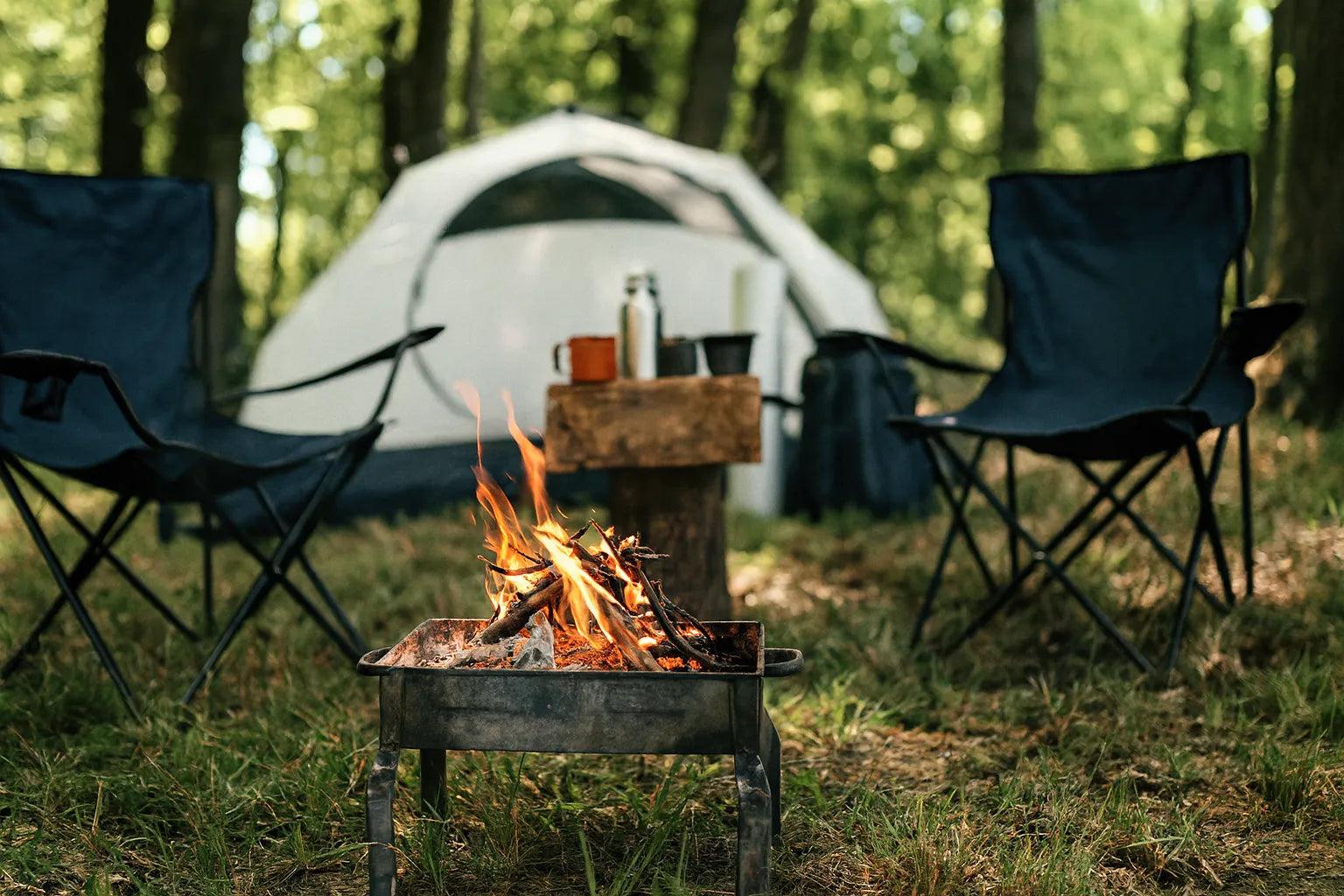 Campement installe en foret avec une tente blanche, un feu de bois dans un brasero et des chaises pliantes autour.