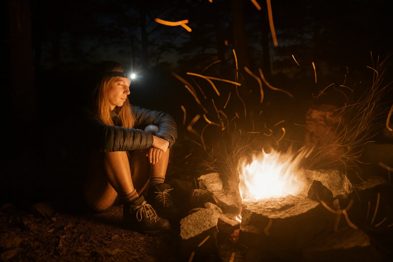 Femme assise près d’un feu de camp avec lampe frontale en forêt.