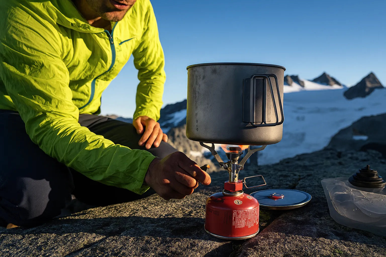 Randonneur allumant un rechaud a gaz portable avec une casserole en aluminium, installe sur un rocher face a un paysage de montagne enneigee.