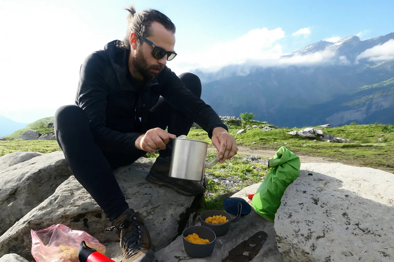 Un randonneur prépare son repas en plein air à l’aide d’une popote en acier posée sur un rocher.