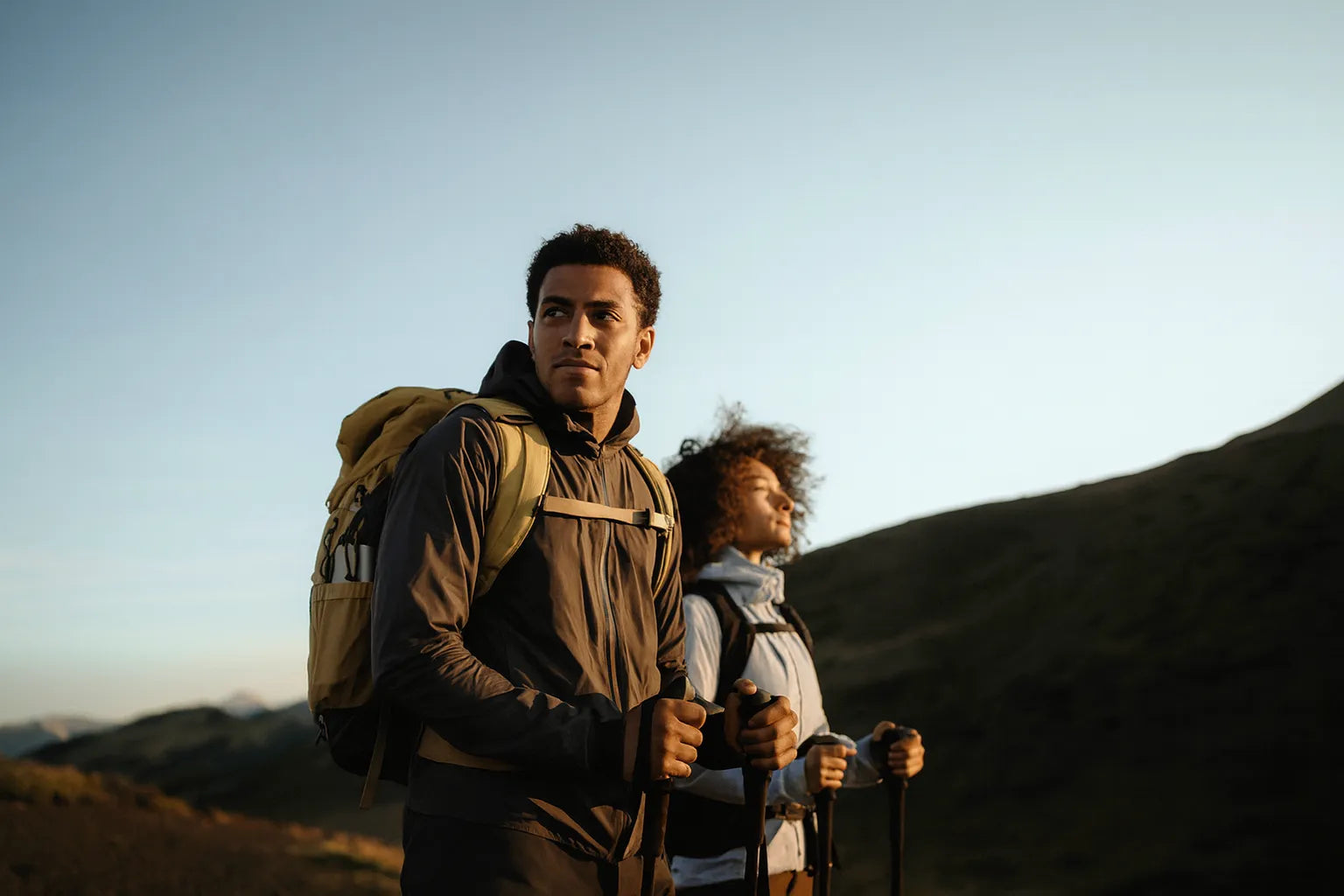 Deux randonneurs equipes de sacs a dos et batons de marche avancent sur un sentier de montagne au lever du soleil.