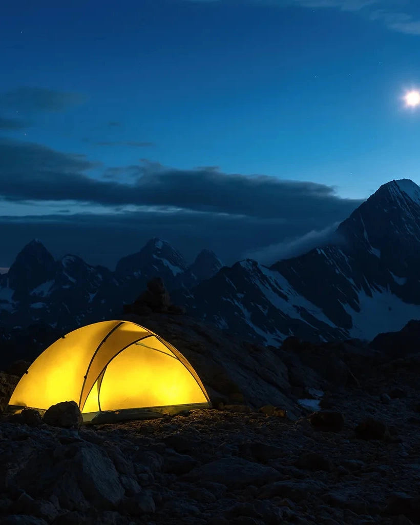 Tente de camping illuminee installee sur un terrain rocheux en haute montagne, sous un ciel nocturne avec la lune brillante et des sommets enneiges en arriere-plan.