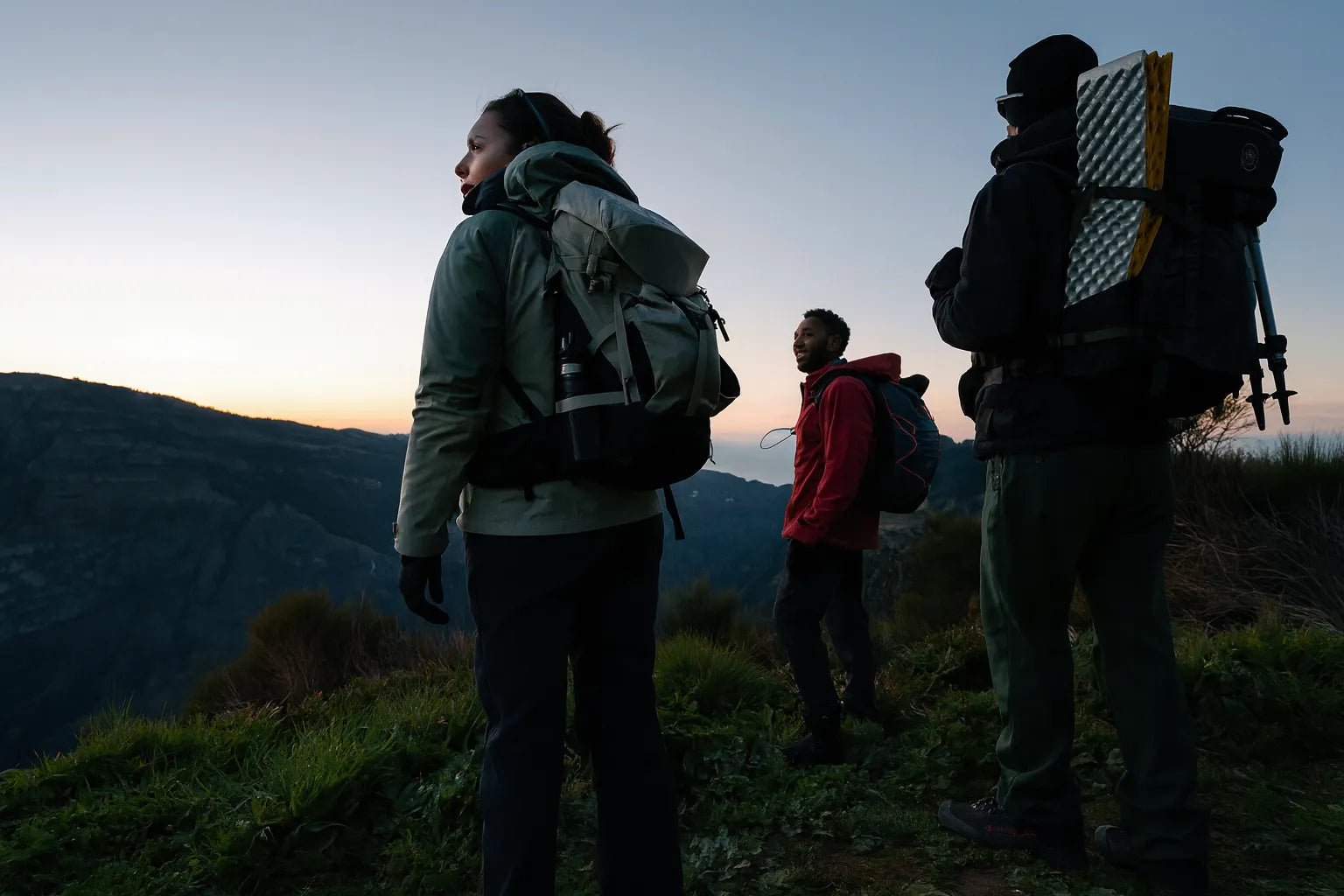 Trois randonneurs equipes de sacs a dos et materiel de bivouac observent le paysage montagneux au crepuscule.