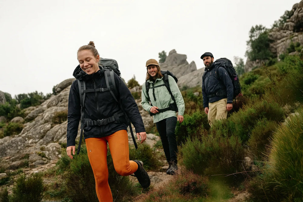 Trois randonneurs avances sur un sentier de montagne rocailleux, equipes de sacs a dos et vetements techniques adaptes aux conditions meteo changeantes.