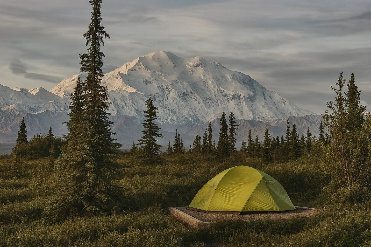 Tente dôme verte pour randonnée hivernale, plantée dans une plaine au pied d’une montagne enneigée.